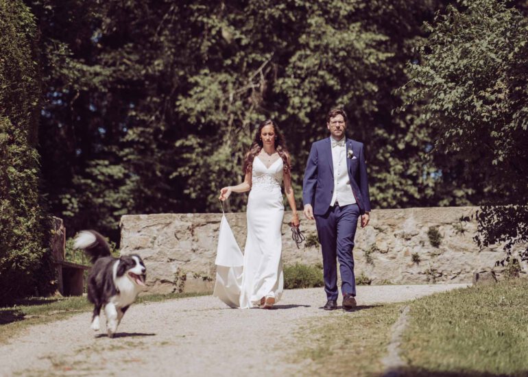 Brautpaar läuft in der prallen Sonne mit ihrem Hund einen Waldweg entlang