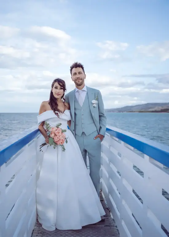 Braut und Bräutigam stehen auf weiß-blauem Steg am Meer und schauen in die Kamera