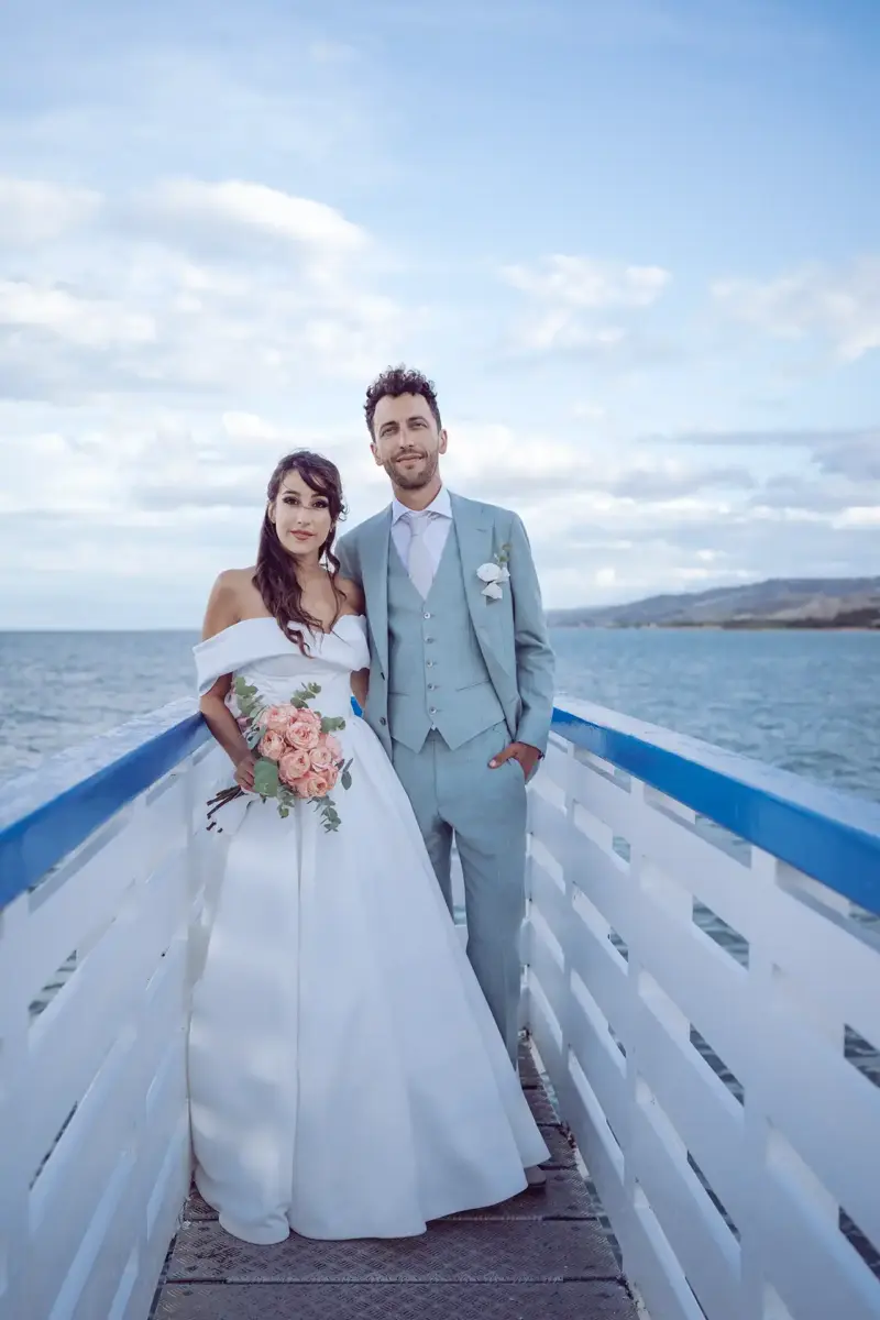 Braut und Bräutigam stehen auf weiß-blauem Steg am Meer und schauen in die Kamera