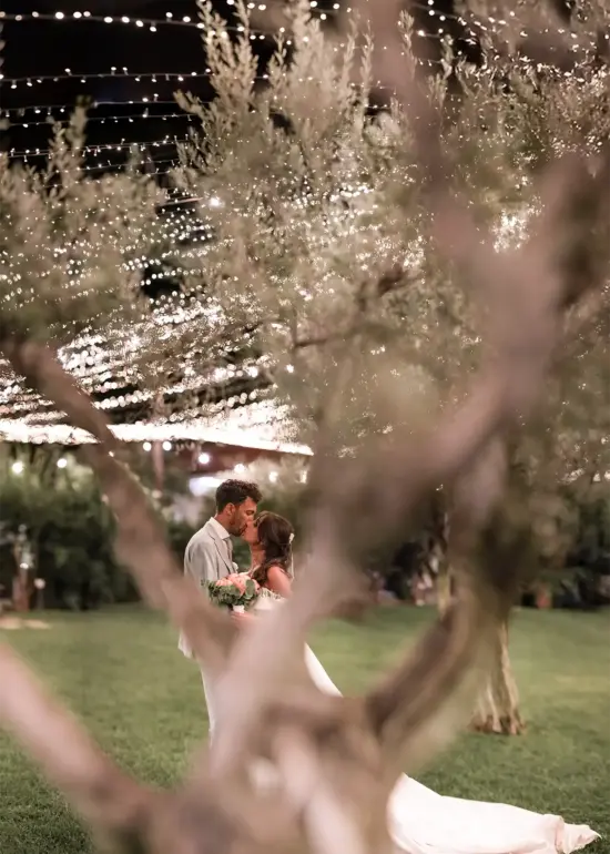 Braut und Bräutigam küssen sich verliebt unter Olivenbäumen und Lichterkette