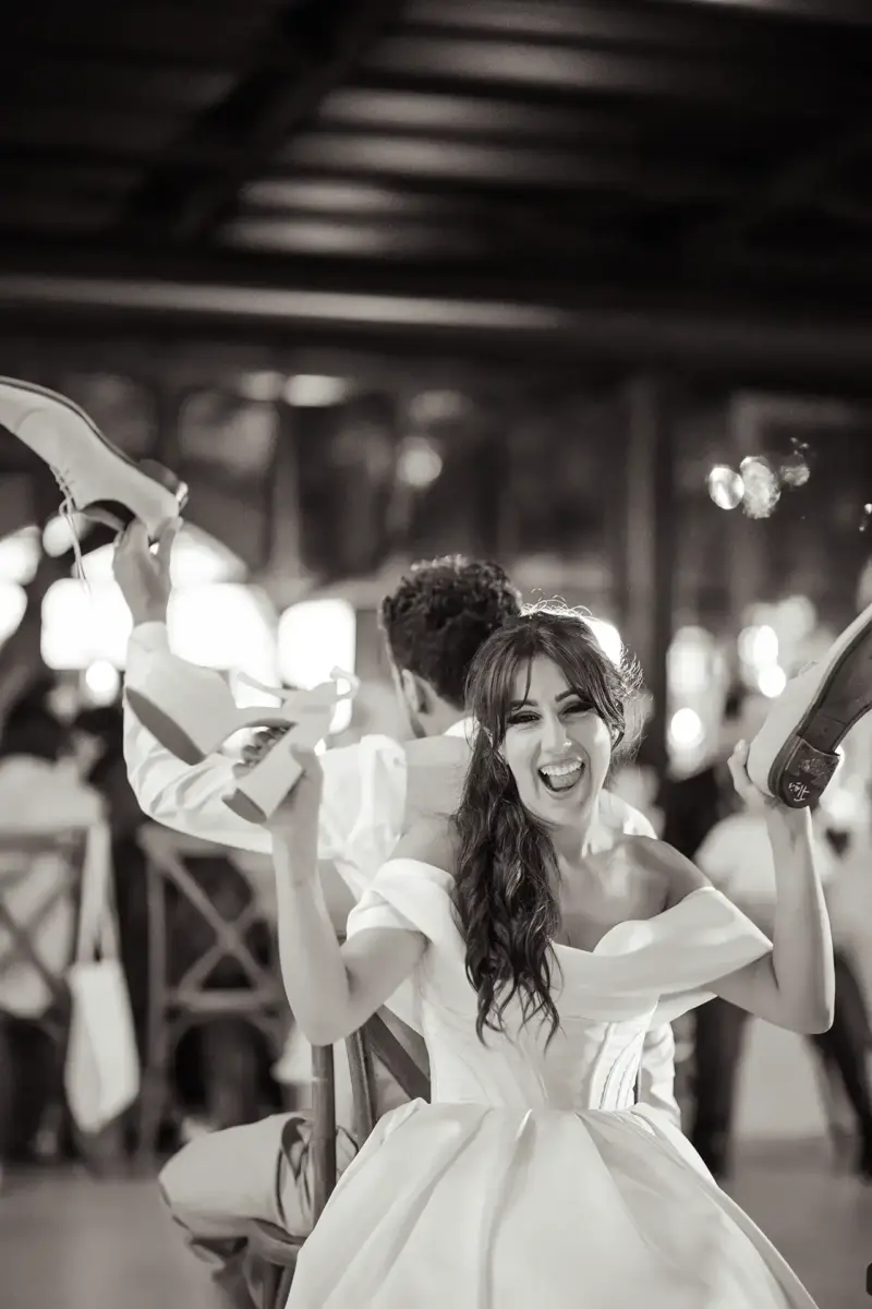 Braut lacht herzlich und hält ihren und den Schuh ihres Mannes in die Höhe, Schwarz-Weiß