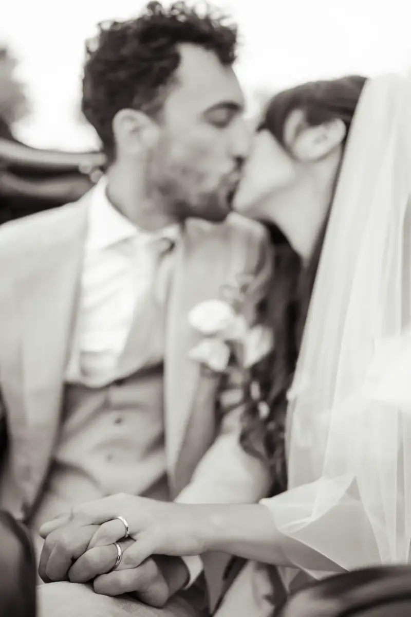 Brautpaar küsst sich auf dem Rücksitz und hält Händchen, sodass man die Eheringe sieht, Schwarz-Weiß