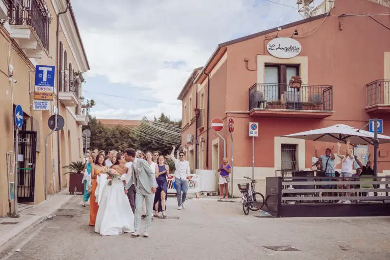 Brautpaar mit allen Hochzeitsgästen im Hintergrund läuft Straße entlang und wird von Passanten gefeiert