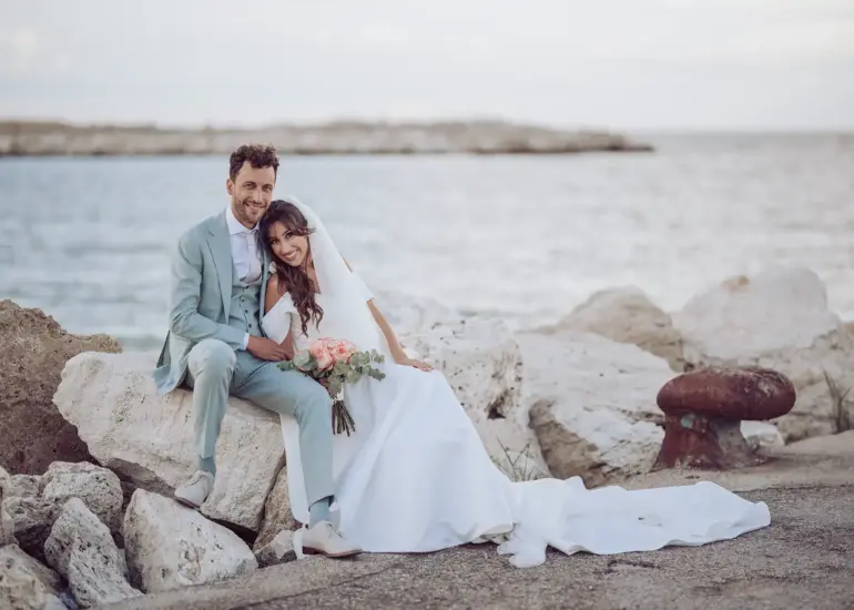 Braut und Bräutigam sitzen am Hafen mit Meer im Hintergrund und schauen in Kamera