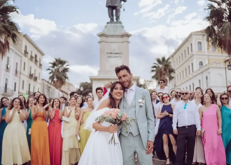 Brautpaar lacht in die Kamera und im Hintergrund sind alle Hochzeitsgäste