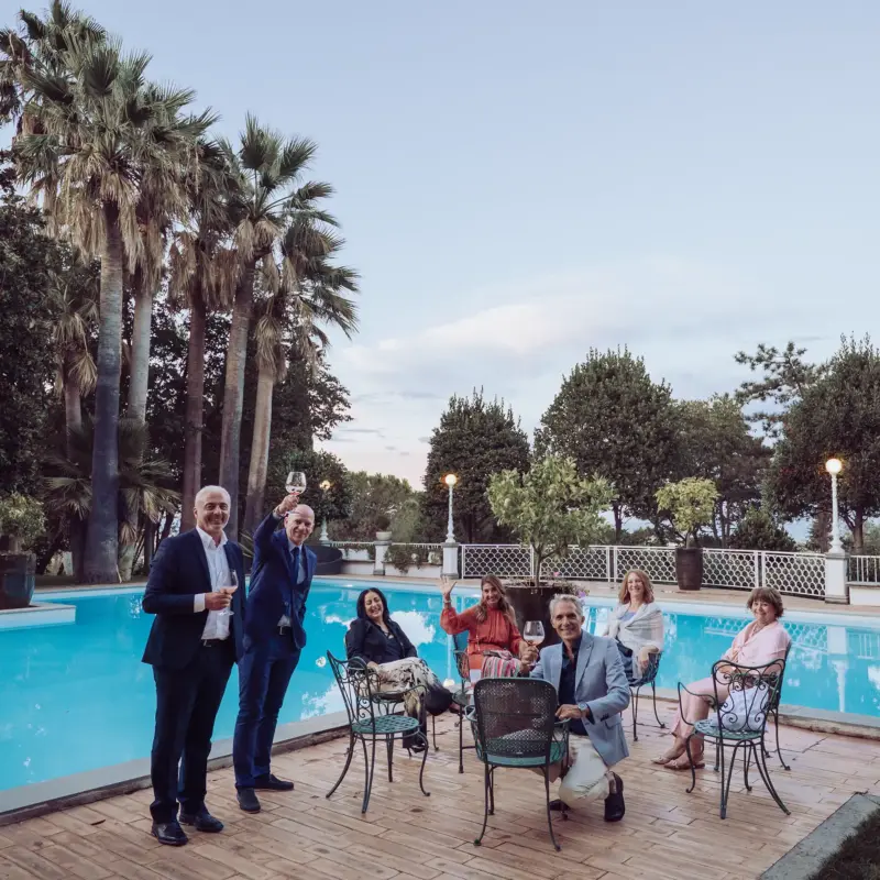 Mehrere Hochzeitsgäste stehen und sitzen vor dem Pool und erheben die Gläser