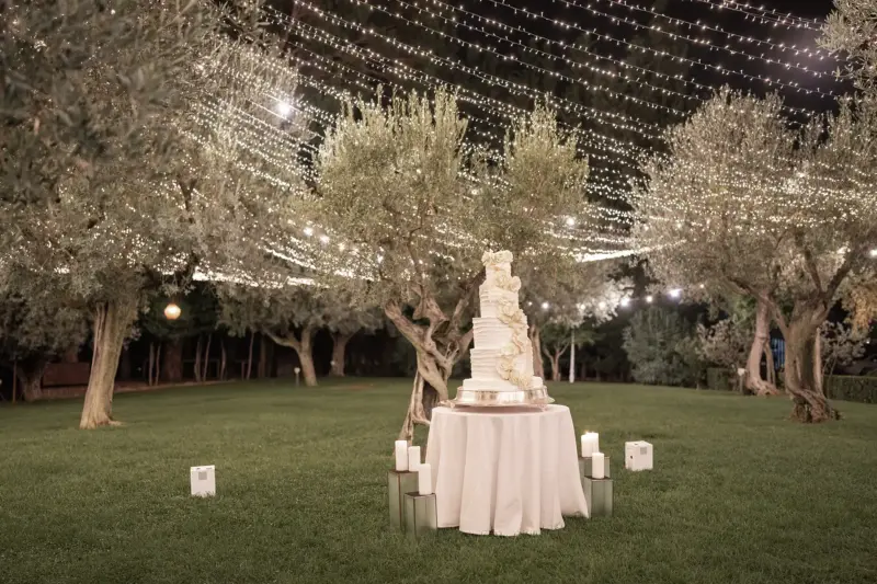 Mehrstöckige Hochzeitstorte steht auf einer Wiese mit Olivenbäumen und vielen Lichterketten