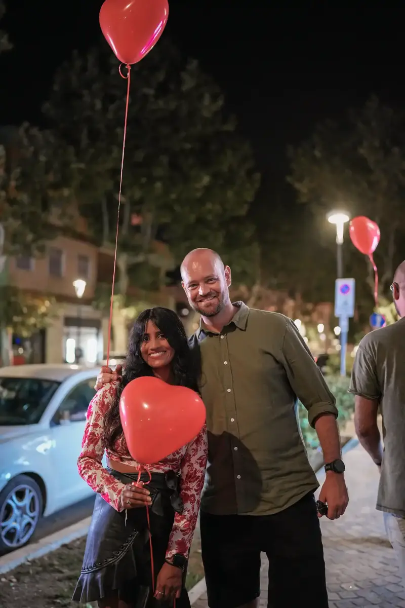 Paar hält zwei Herzluftballons in den Händen und lacht in die Kamera