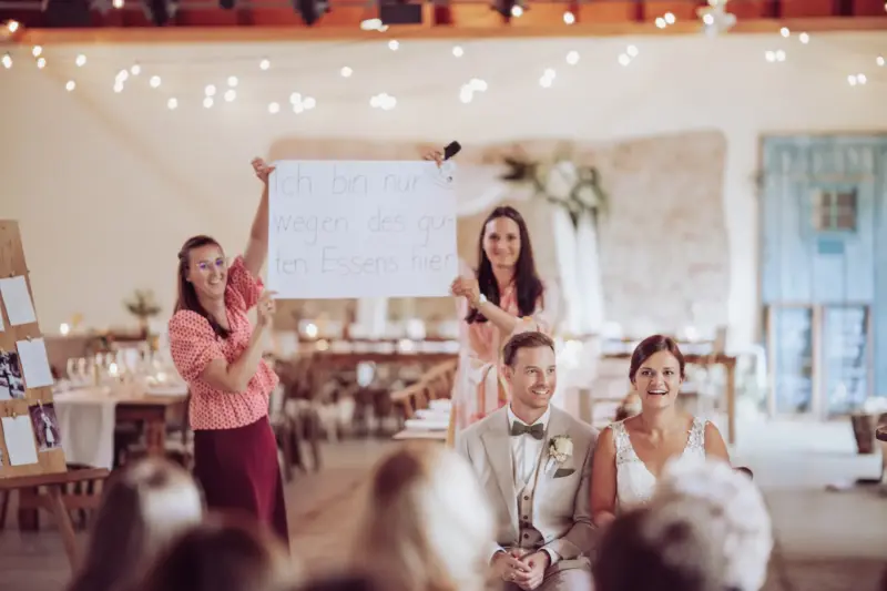 Brautpaar sitzt vor den Hochzeitsgästen, im Hintergrund wird ein Schild hochgehalten